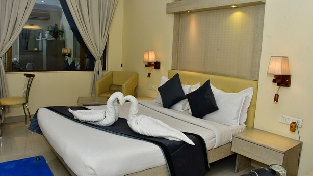 A hotel room with a bed, two bedside tables, a chair, a desk, and a window with curtains. - THE RASS HOTEL (Kanpur)