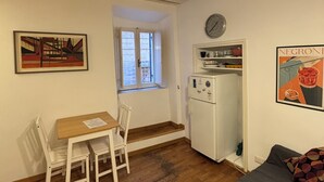 Ett litet rum med trägolv, ett litet bord med stolar, en säng med ett mönstrat överkast, ett kylskåp, en klocka och två inramade tavlor på väggarna.
