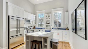 A modern kitchen with white cabinetry, a stainless steel refrigerator, a central island with stools, and a view of the outside through large windows. - River Park Retreat! Walk to River, Trails & Town Life (Buena Vista)