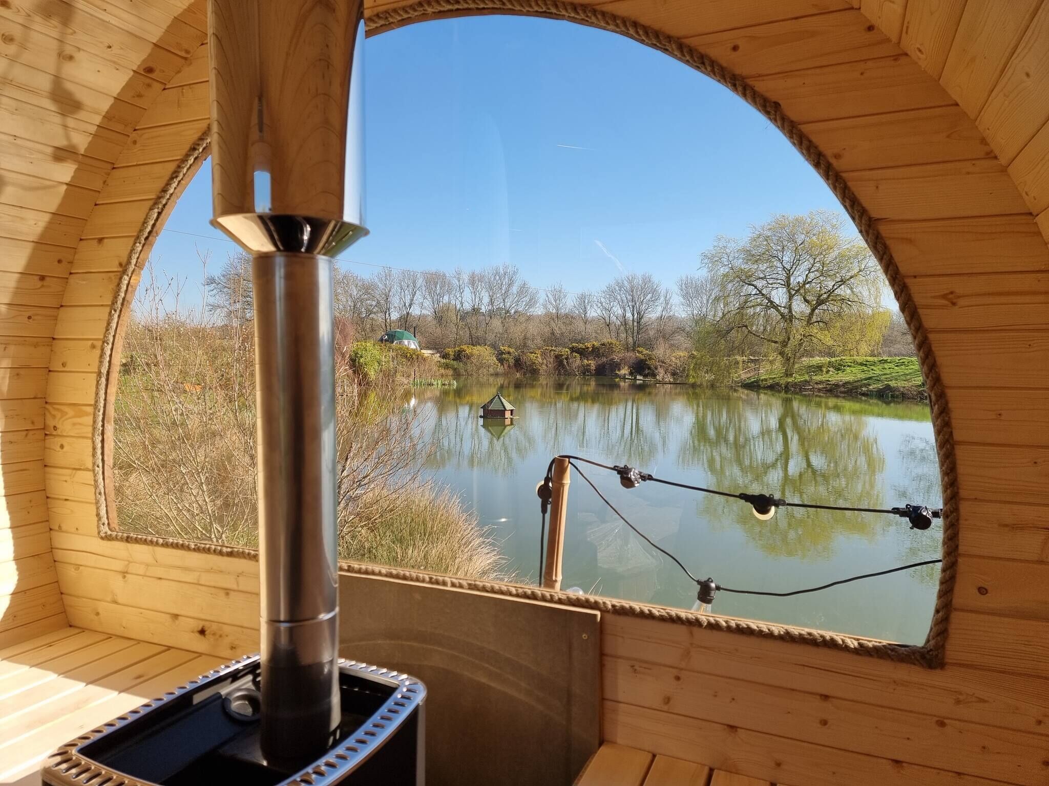 A wooden cabin with a large arched window overlooking a calm lake, a chimney, and a fishing rod.