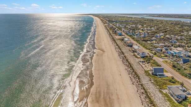 Vista aérea de uma área costeira com uma extensa praia de areia, uma fileira de casas e uma ponte ao longe.