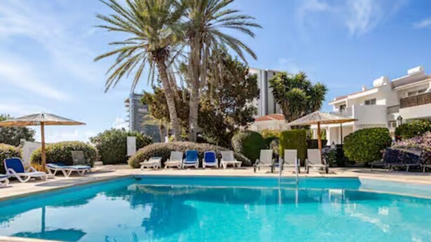 A swimming pool with lounge chairs and umbrellas, surrounded by palm trees and a building. - Gorgeous Apartment in Paradise Complex (Paphos)