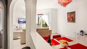 A modern living room with a large window, a sofa, and a dining area with a red tablecloth. - Gorgeous Apartment in Paradise Complex (Paphos)