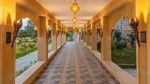 A corridor with patterned flooring, wall-mounted lights, and a view of greenery outside. - Raj Leela Heritage Resort by Estherea (Ghanerao)