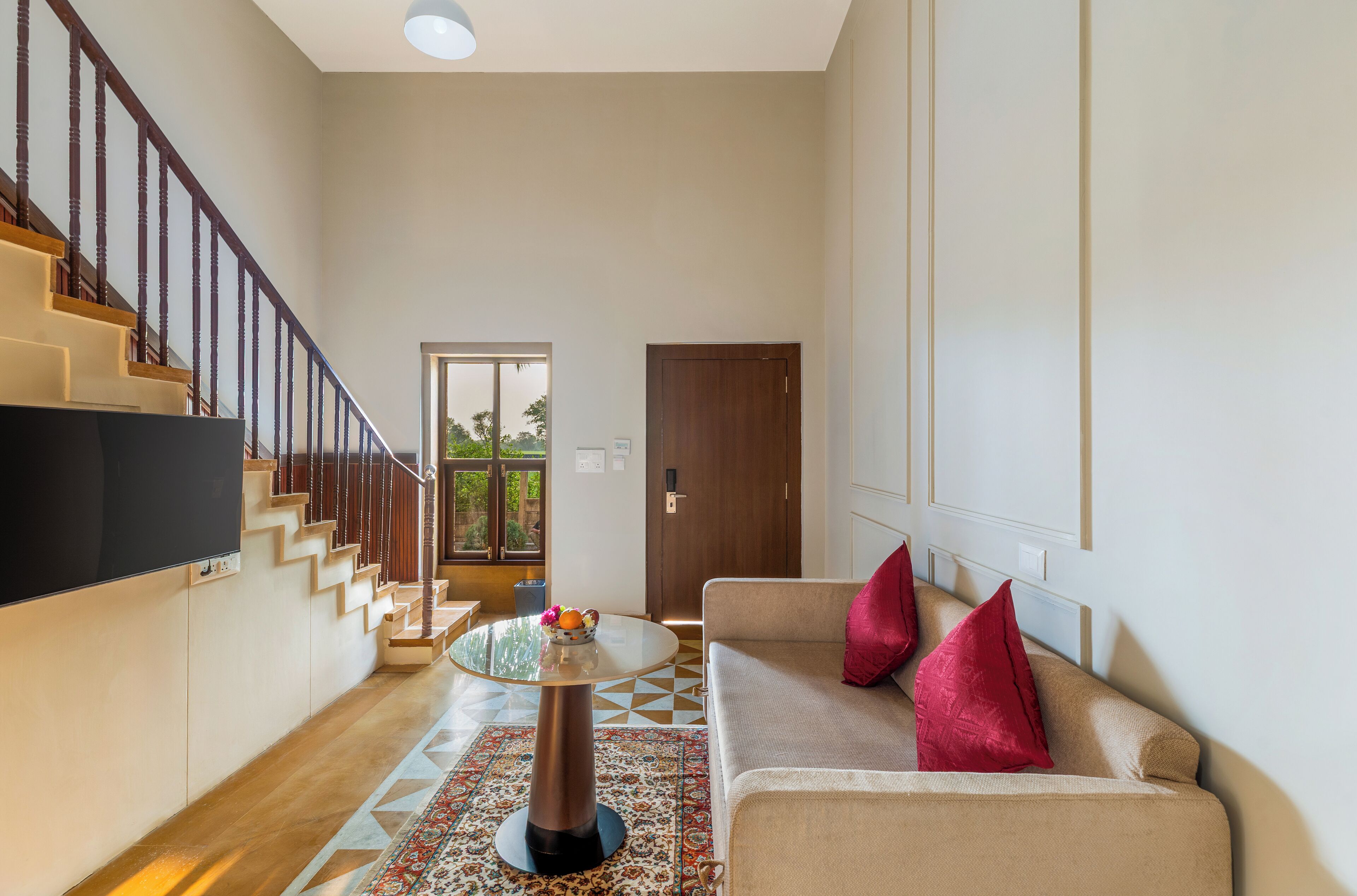 Una sala de estar moderna con una escalera, un sofá con cojines rojos, una mesa redonda con fruta y un televisor montado en la pared.