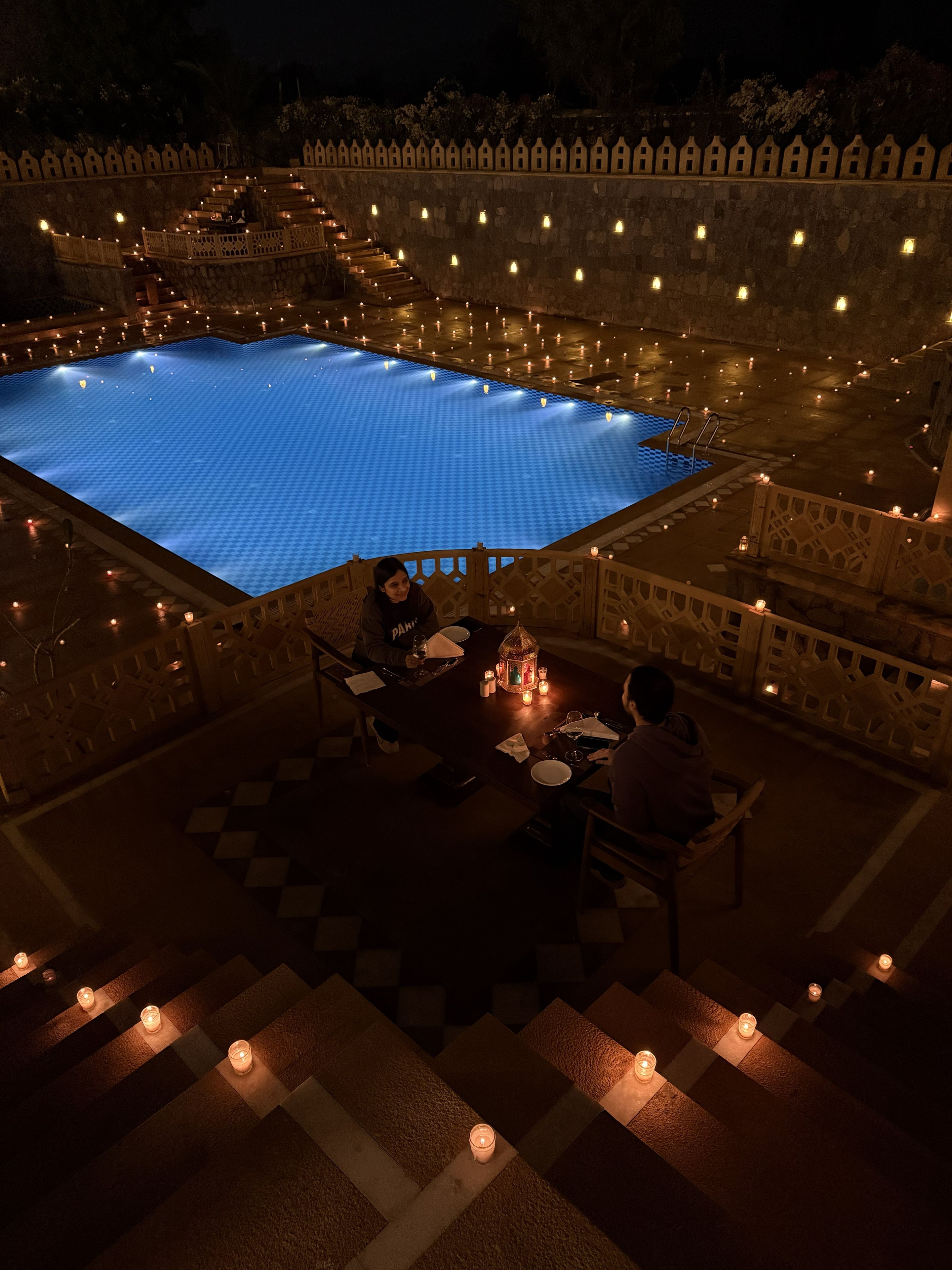 Zona de piscina con una mesa y sillas, rodeada por un muro de piedra con luces distribuidas uniformemente.
