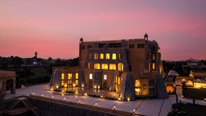 A historic building with multiple levels and arched windows, surrounded by a well-lit courtyard and a stone wall. - Raj Leela Heritage Resort by Estherea (Ghanerao)