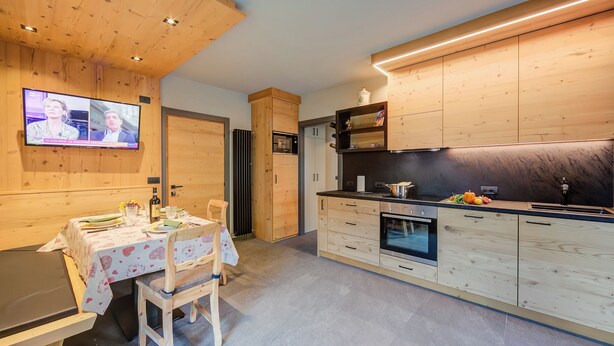 A modern kitchen with wooden cabinetry, a dining area with a table set for two, and a television mounted on the wall. - Loredana 2 (Livigno)