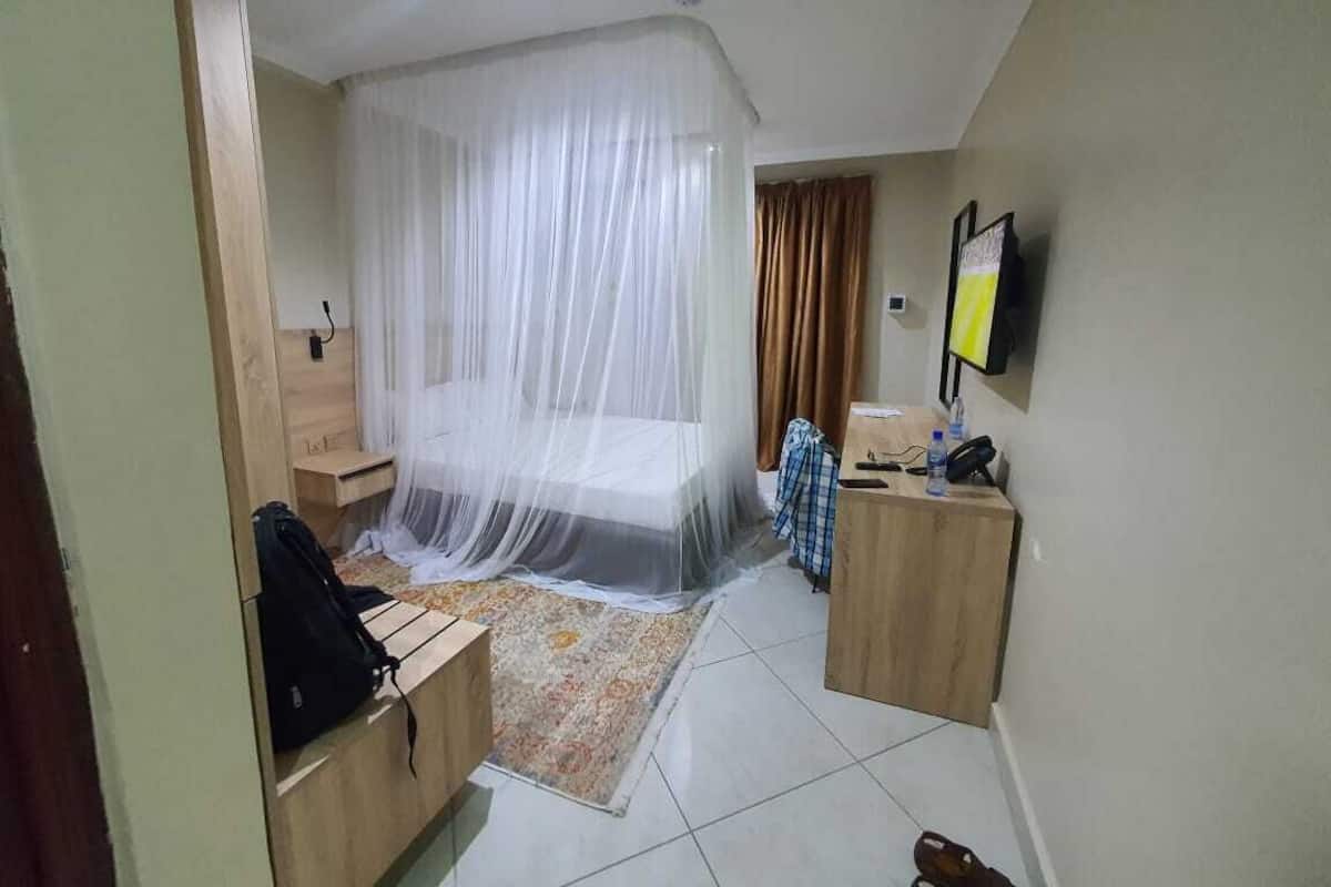 A hotel room with a bed covered by a mosquito net, a wooden desk, a chair, a small table, a television mounted on the wall, and a tiled floor.