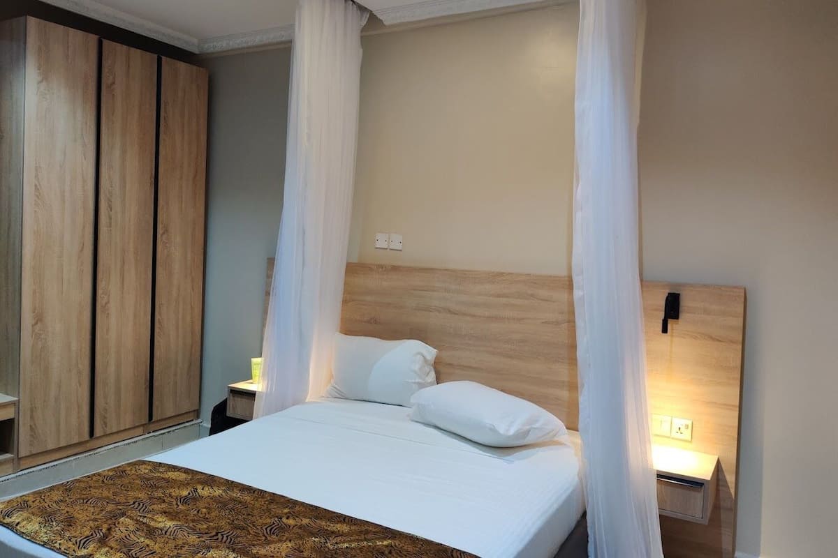 A bedroom with a bed, white curtains, a wooden headboard, a bedside table with a lamp, and a nightstand.