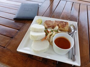 A white plate with a grilled fish, white rice, a small salad, and a bowl of sauce on a wooden table.