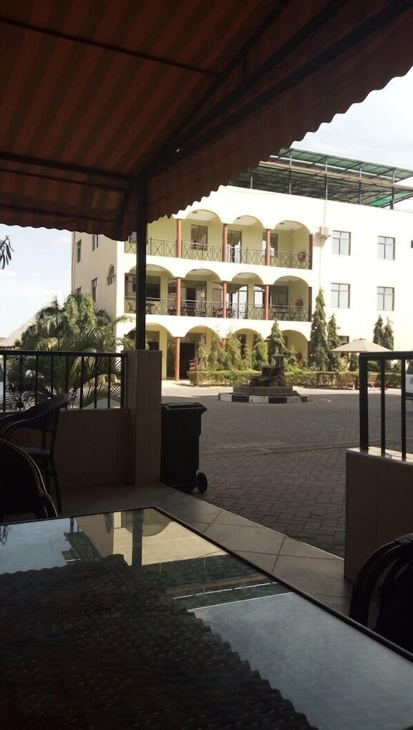 Un espacio al aire libre techado con mesa de billar, un cubo de basura y una fuente. Al fondo, un edificio de varios pisos con balcones y un jardín.