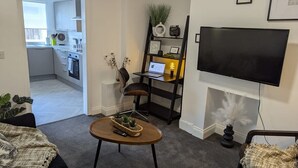 A living room with a sofa, a coffee table, a TV, a bookshelf, and a kitchen in the background. - 4 BR House | Unli Parking | Bathtub (West Midlands)