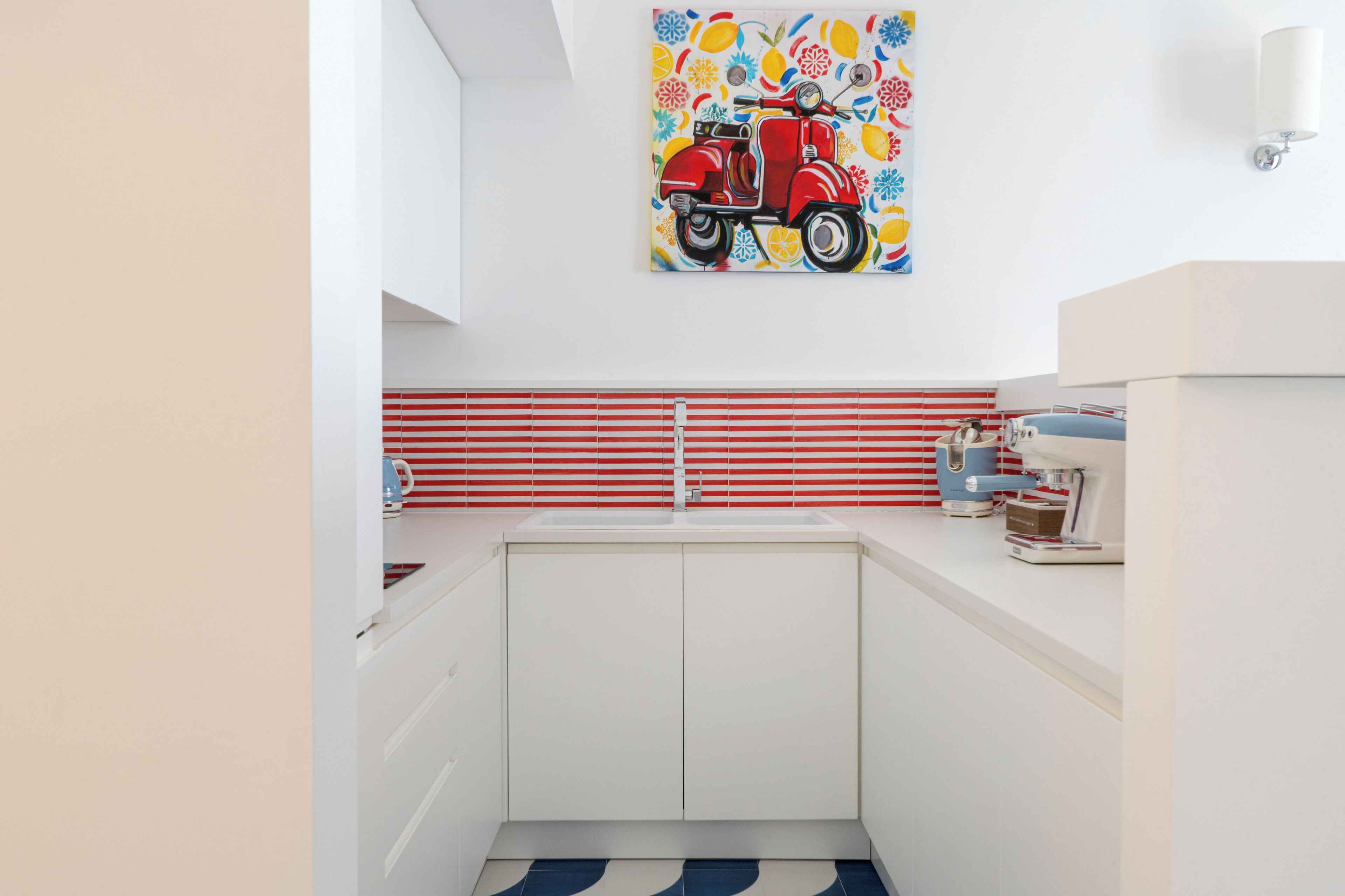 A modern kitchen with white cabinets, a red and white striped backsplash, and a colorful painting of a red scooter.