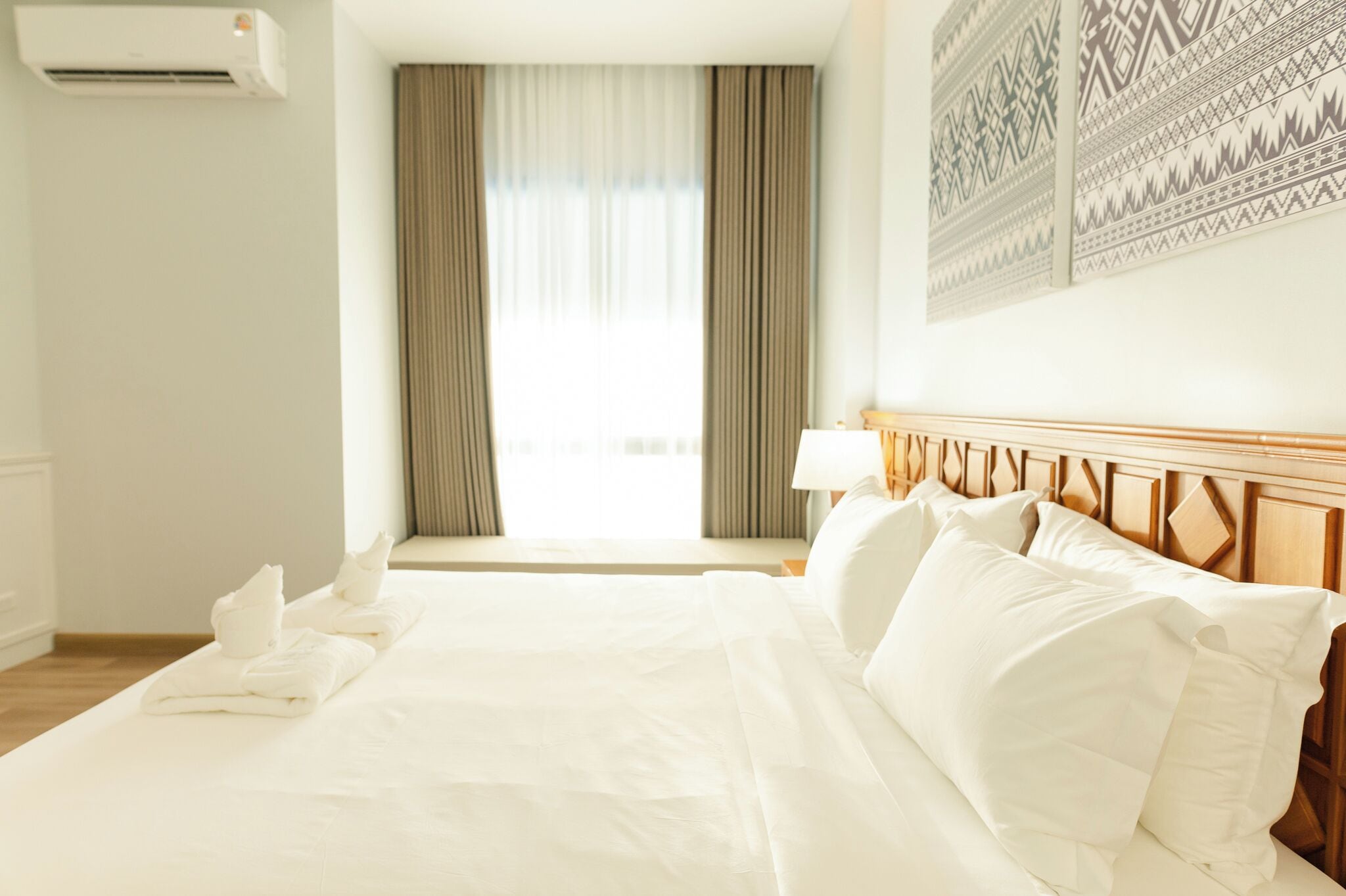 A hotel room with a large bed, a window with curtains, an air conditioning unit, and a wall with patterned artwork.
