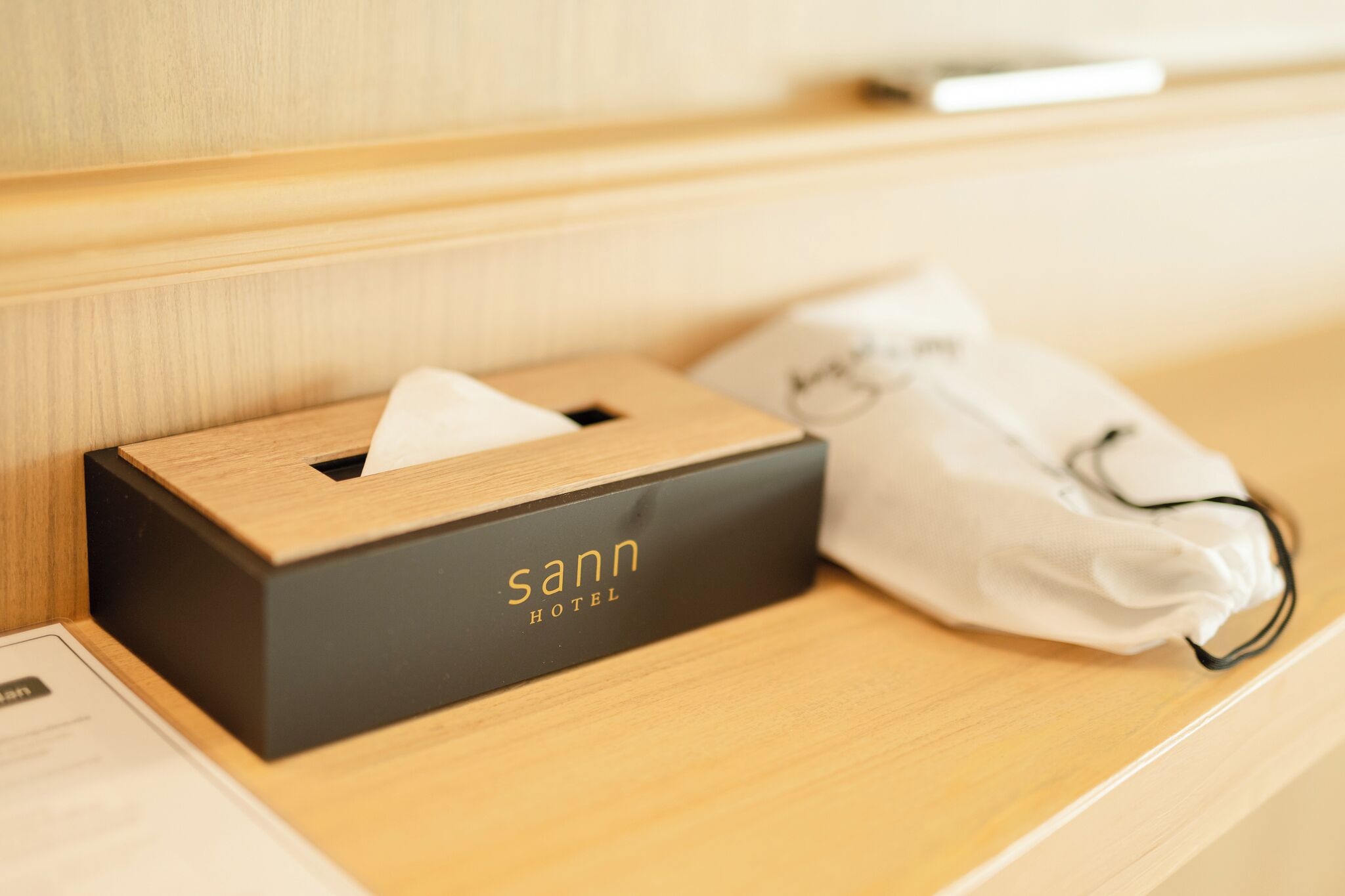 Ein Hotelzimmer mit einem Nachttisch aus Holz, einer Taschentuchbox mit dem Aufdruck 'Sann Hotel' und einer weißen Gesichtsmaske.