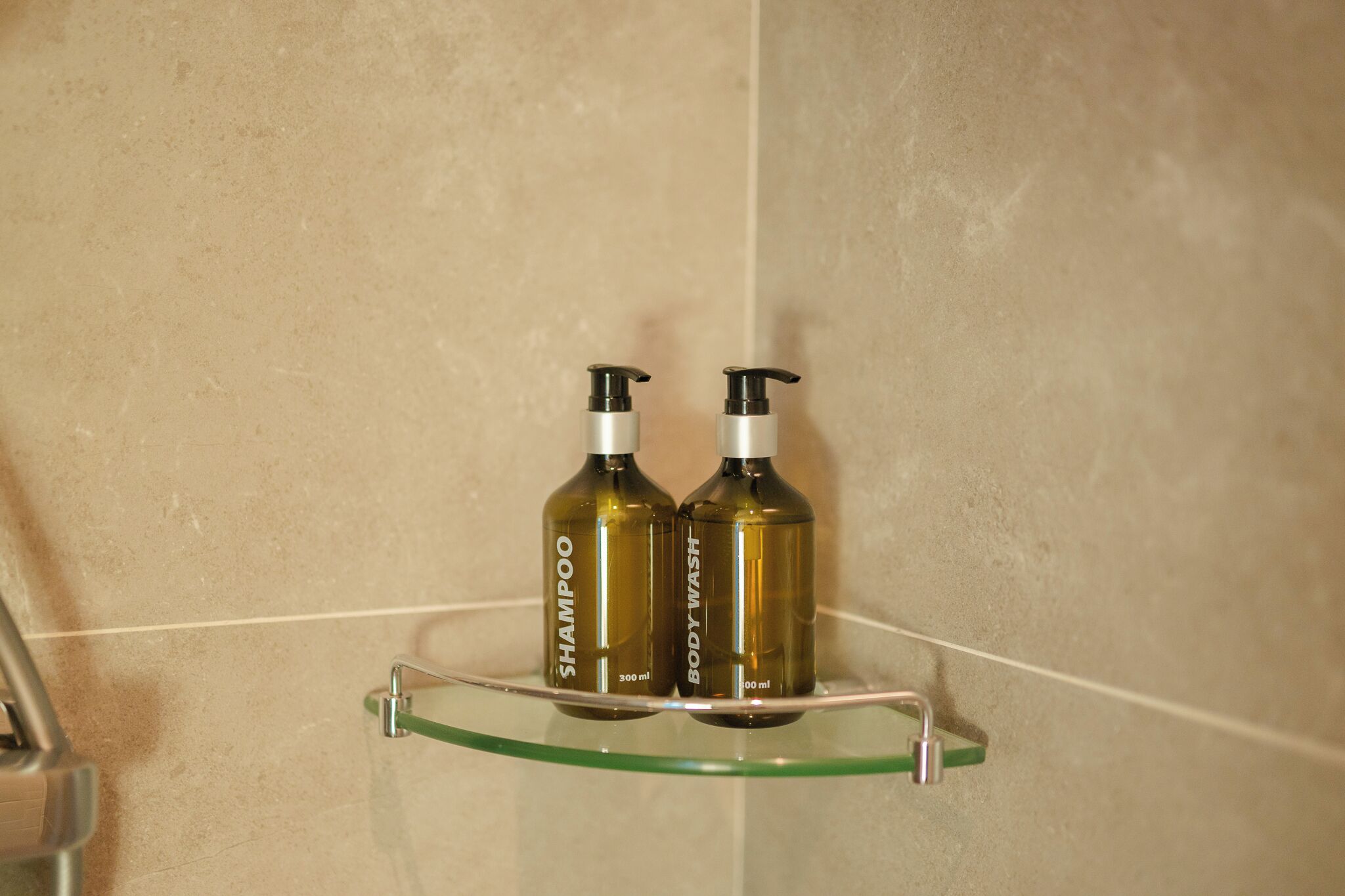 Dos botellas de champú y gel de ducha sobre un estante de vidrio en un baño con azulejos color beige.