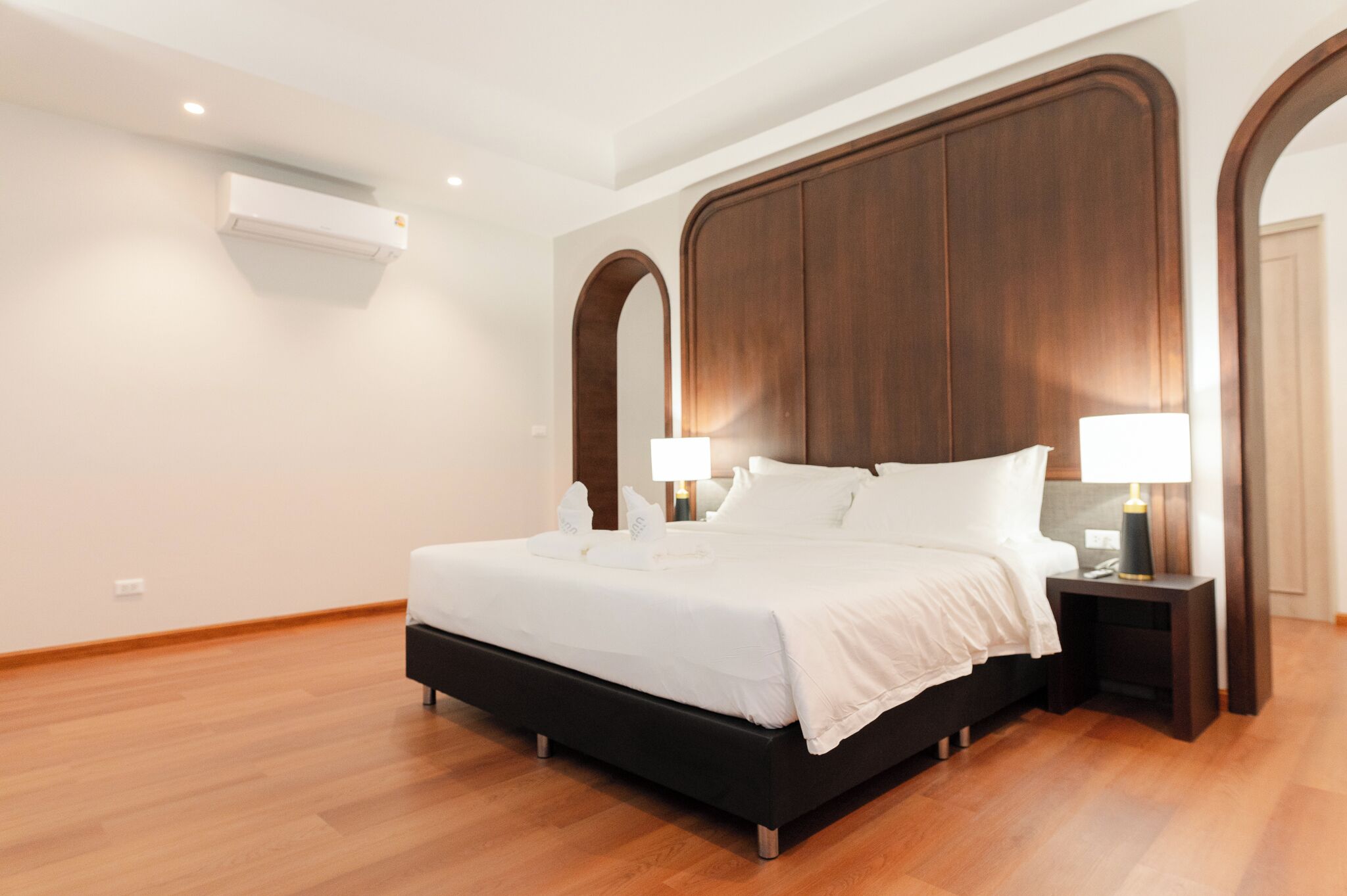 Quarto de hotel moderno com uma cama grande, cabeceira de madeira, duas mesas de cabeceira com luminárias e um aparelho de ar condicionado na parede.