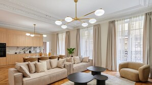Una sala moderna con un sofá seccional, una mesa de centro y un sillón. Hay una zona de cocina con encimeras de mármol y armarios de madera. La habitación tiene ventanas amplias con cortinas y un ventilador de techo con múltiples luces.