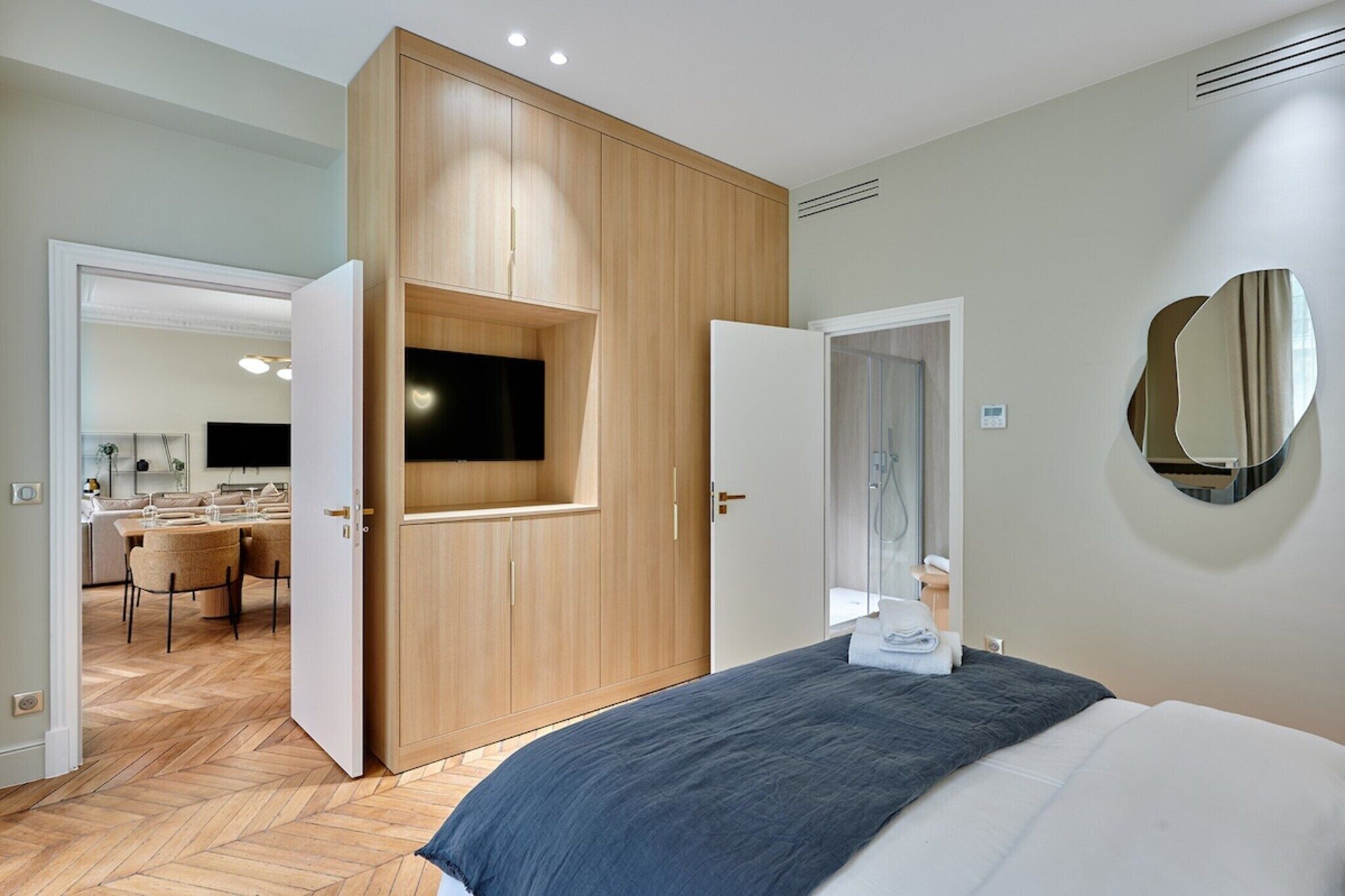 A modern hotel room with a bed, a built-in TV, a mirror, and a wooden wardrobe.