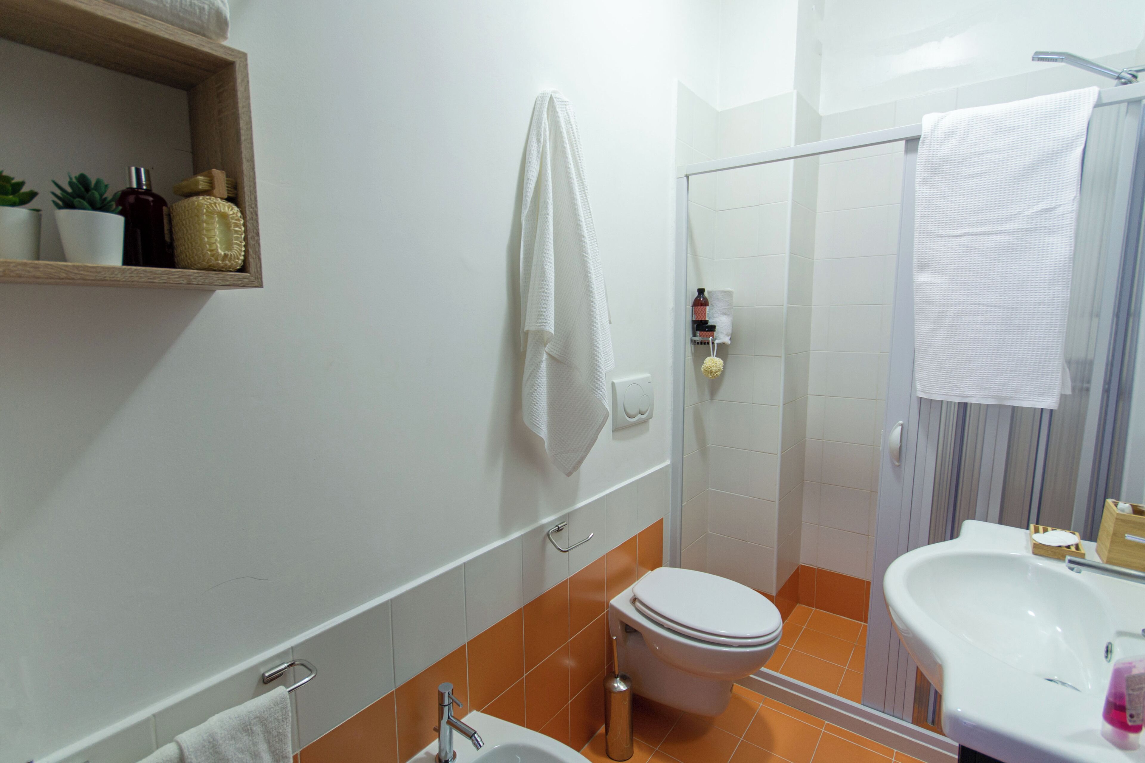 A bathroom with a toilet, sink, and shower area. There are towels hanging on the wall and a shelf with toiletries.