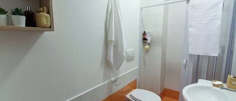 Baño con inodoro, lavabo y zona de ducha. Hay toallas colgadas en la pared y un estante con artículos de aseo.