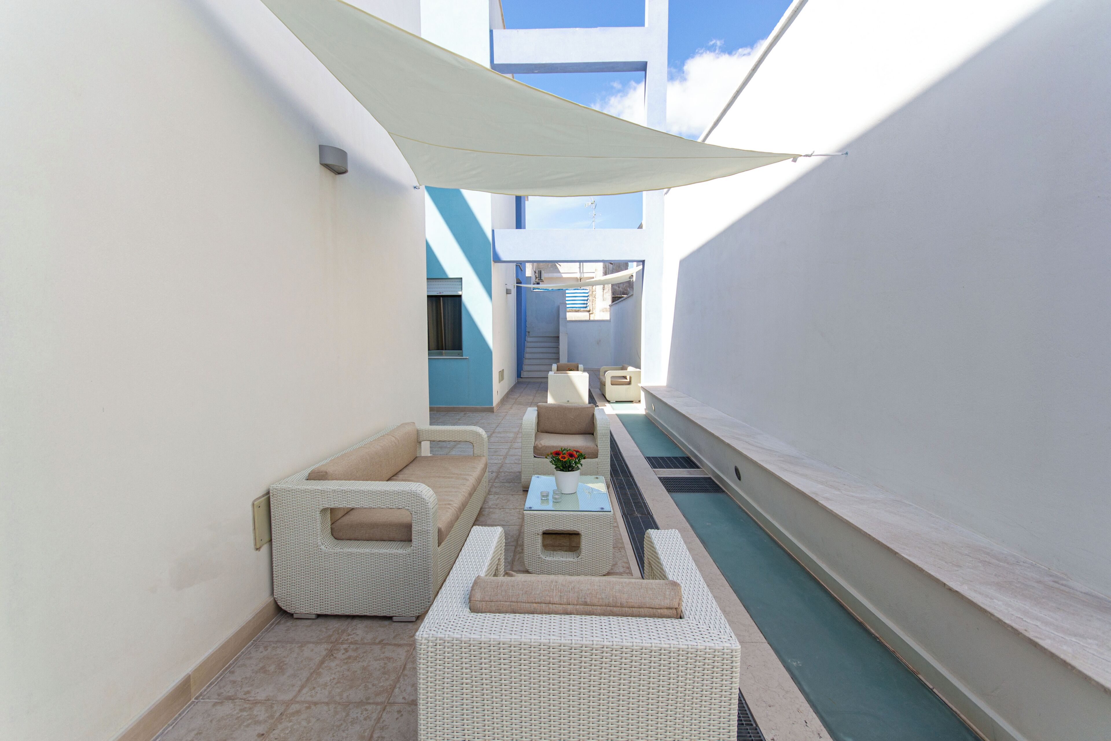 A narrow outdoor corridor with a pool, white wicker furniture, and a staircase leading to an upper level.