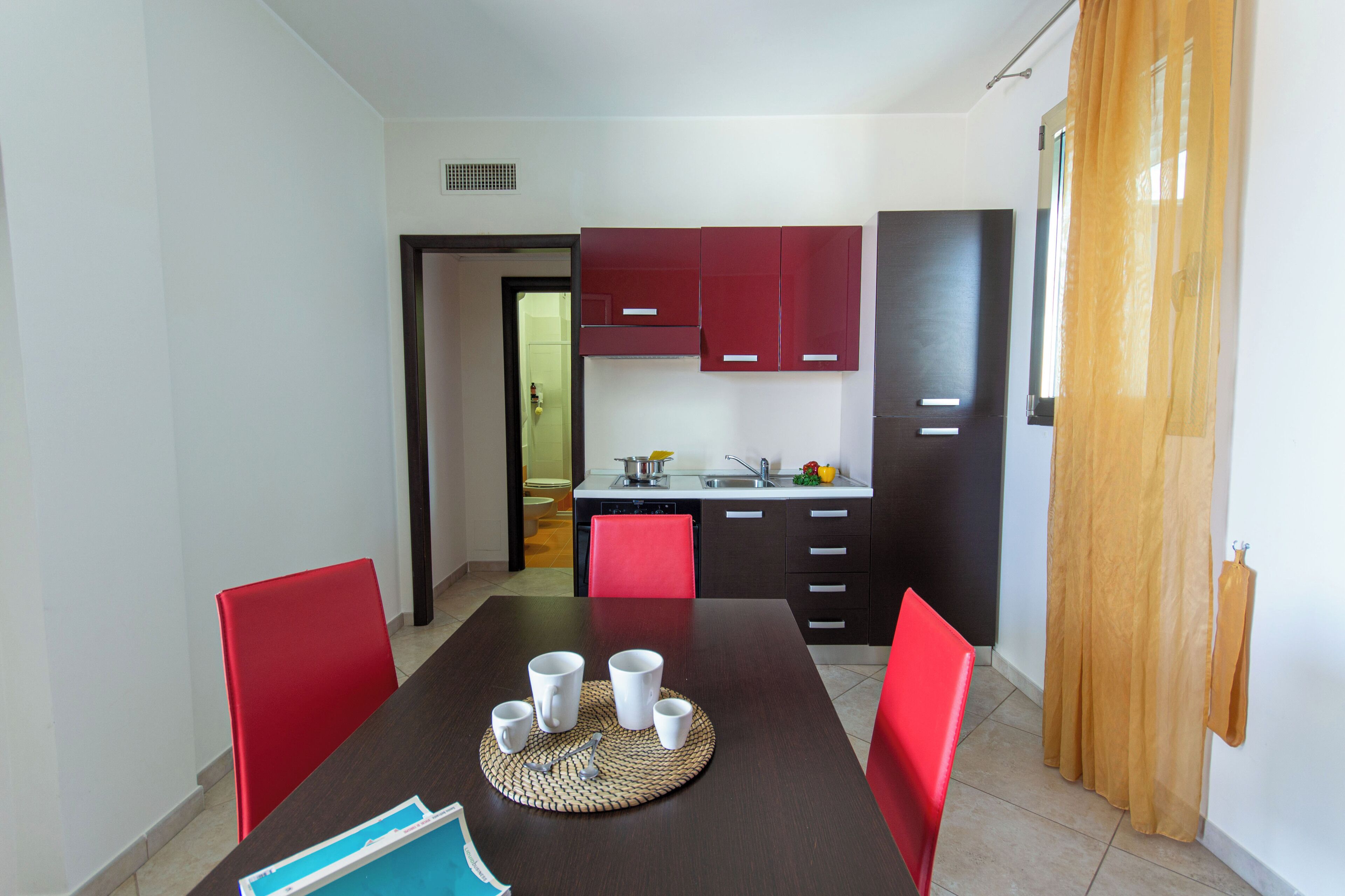 Un coin repas avec une table dressée pour quatre personnes, des chaises rouges et une cuisine en arrière-plan.