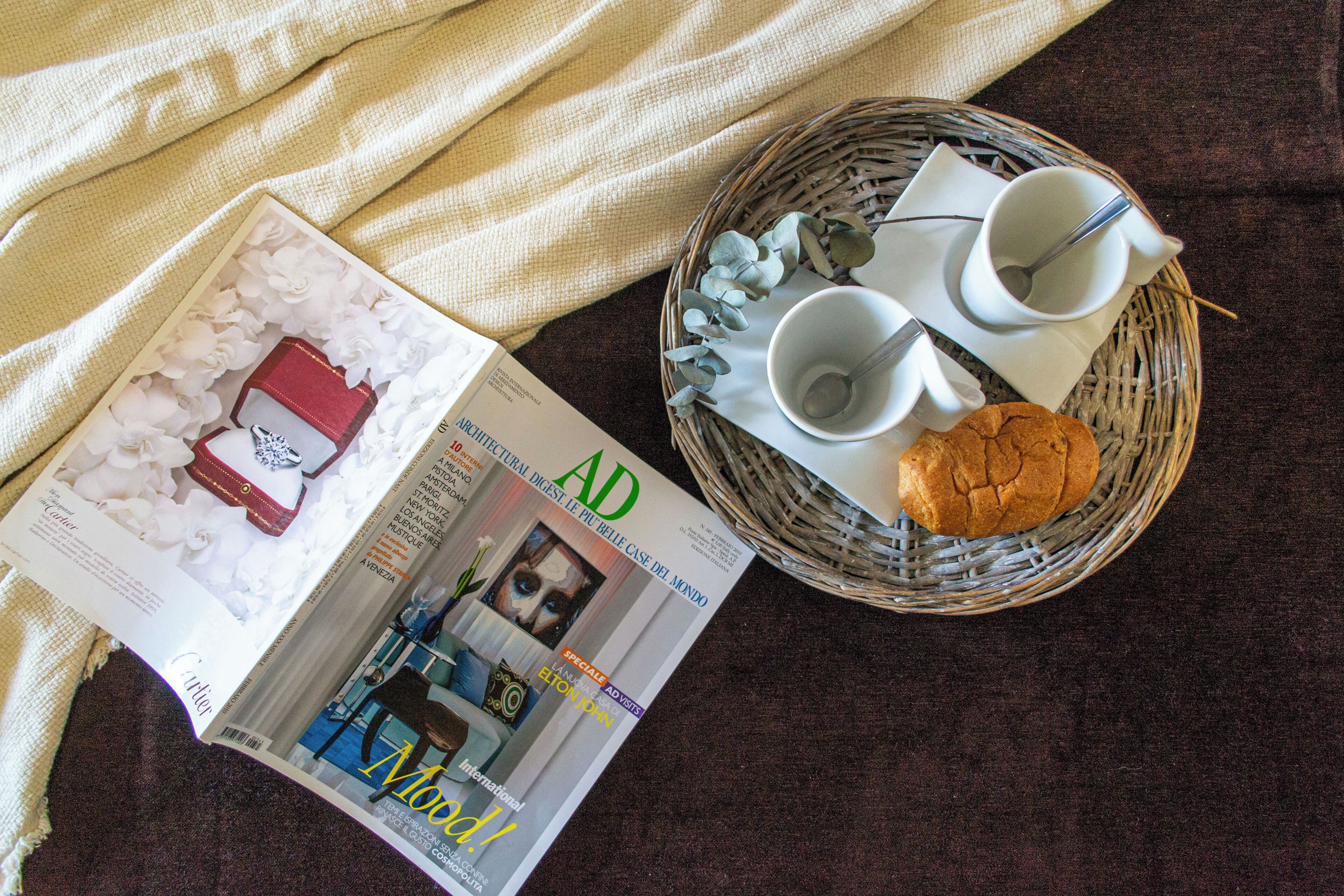A magazine on a table with a wicker basket containing a cookie and two white cups.