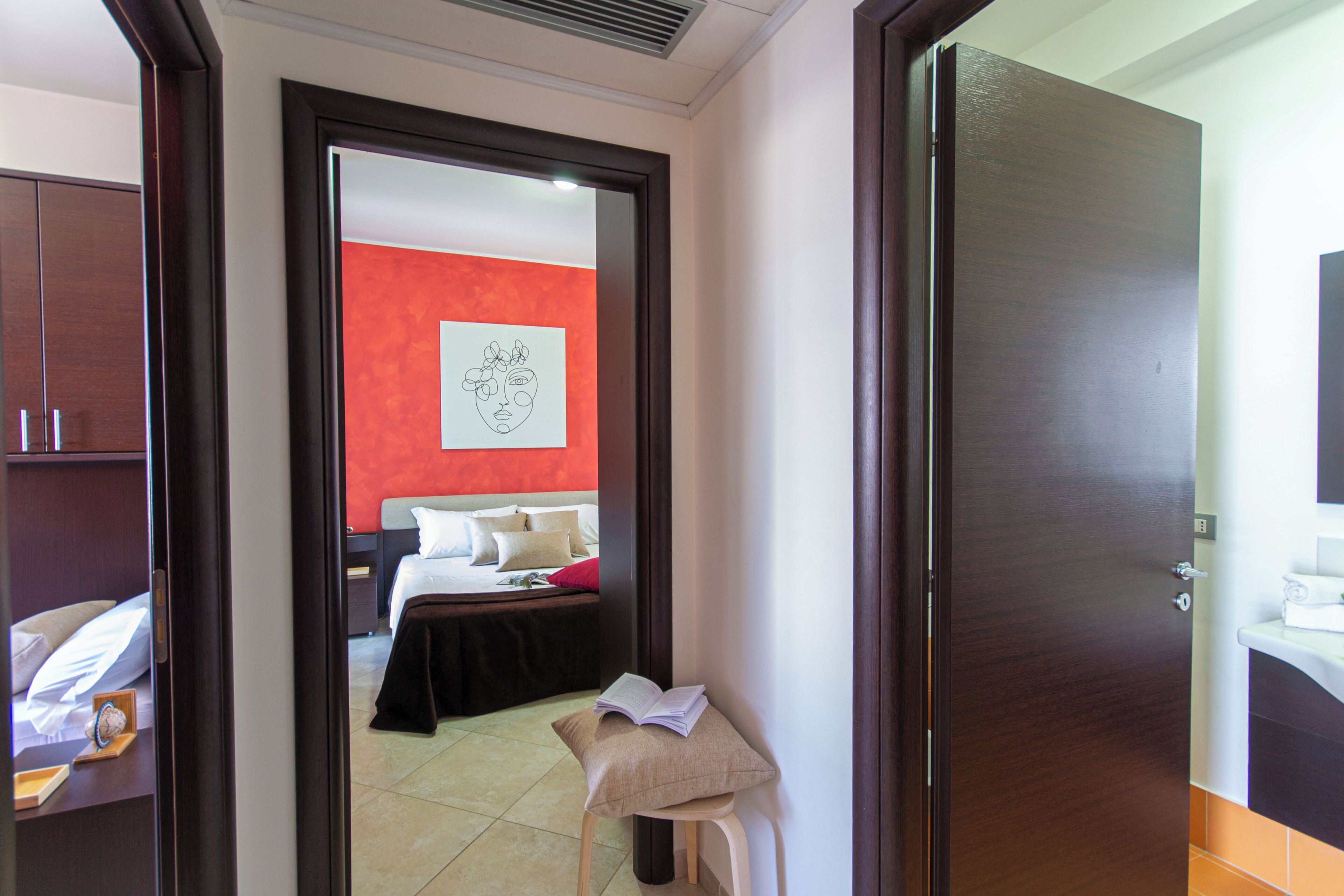 A hotel room with a bed, a bedside table with a book, a small stool, and a red accent wall.