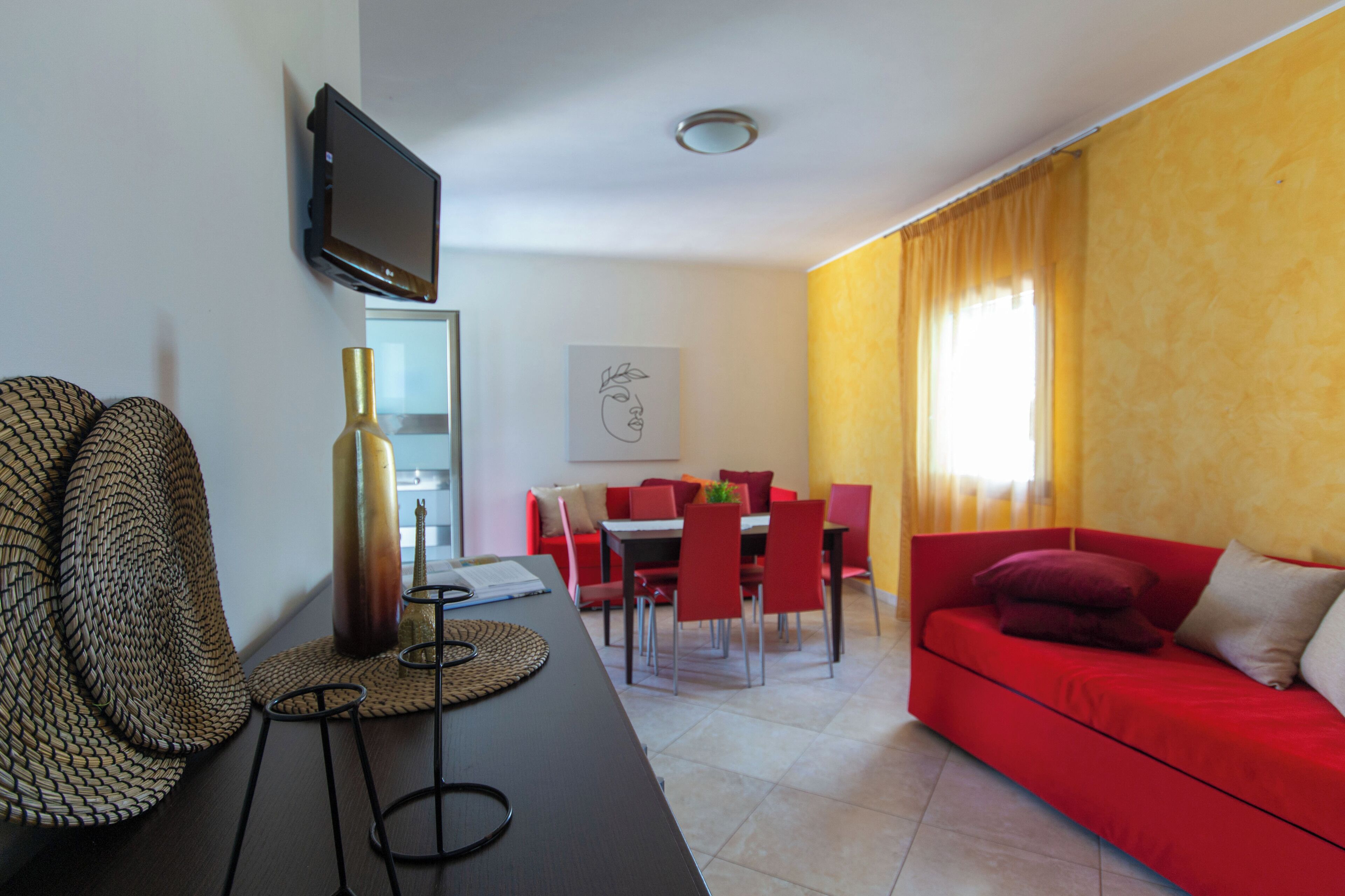 Un salon moderne avec un canapé rouge, un coin repas avec des chaises rouges, un téléviseur à écran plat fixé au mur et une grande fenêtre ornée de rideaux jaunes.