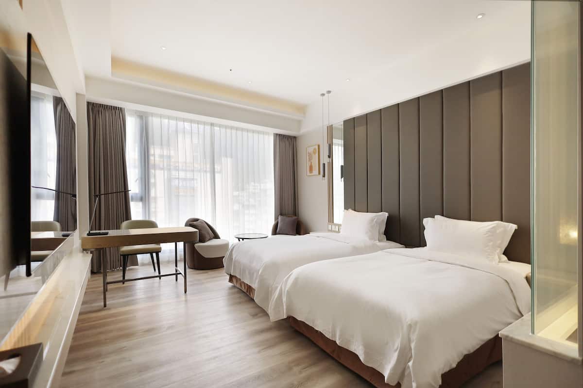 Una habitación de hotel moderna con dos camas, un escritorio y un amplio ventanal con cortinas.