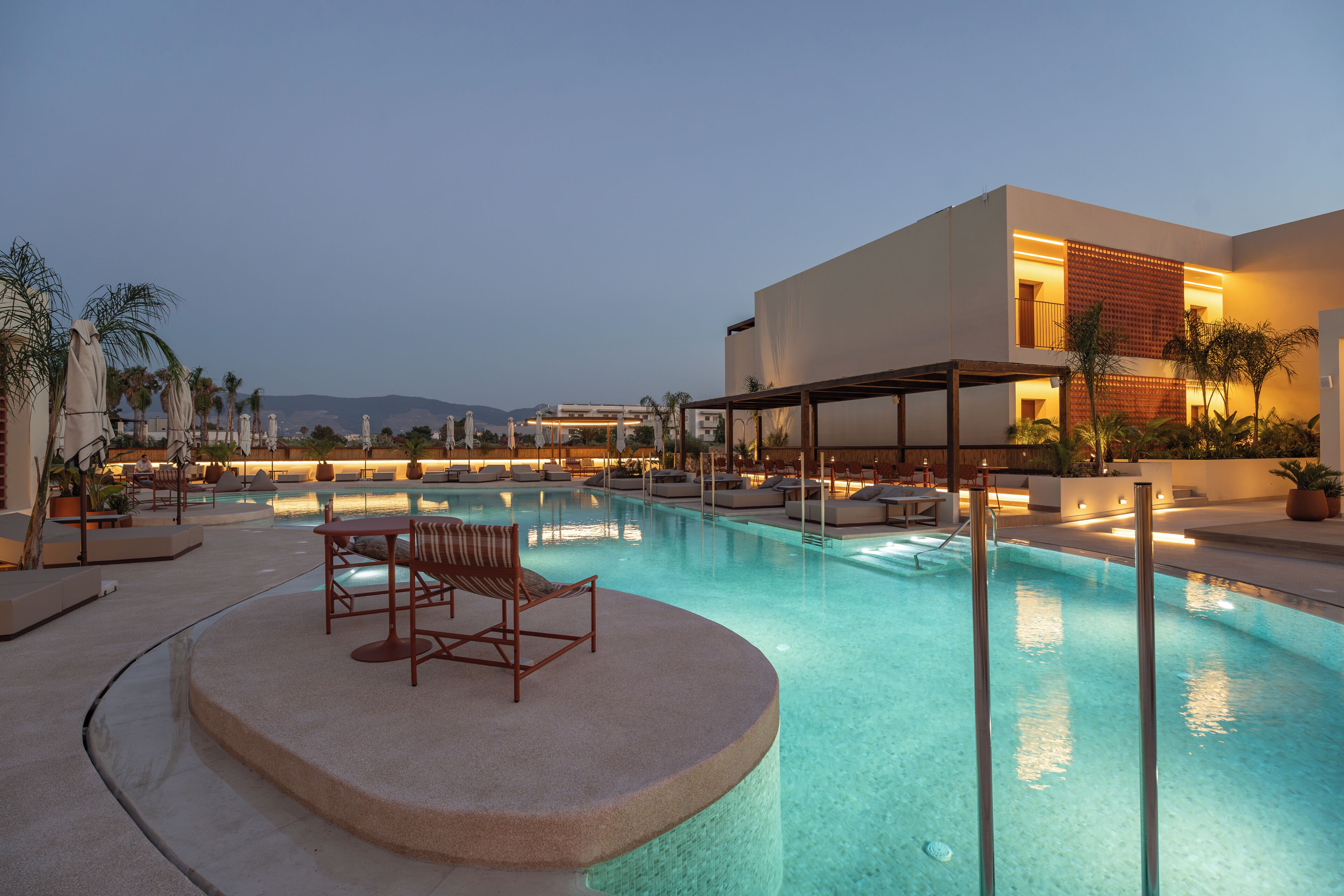 Una piscina con sillones de descanso, un espacio de estar cubierto y un edificio moderno al fondo.