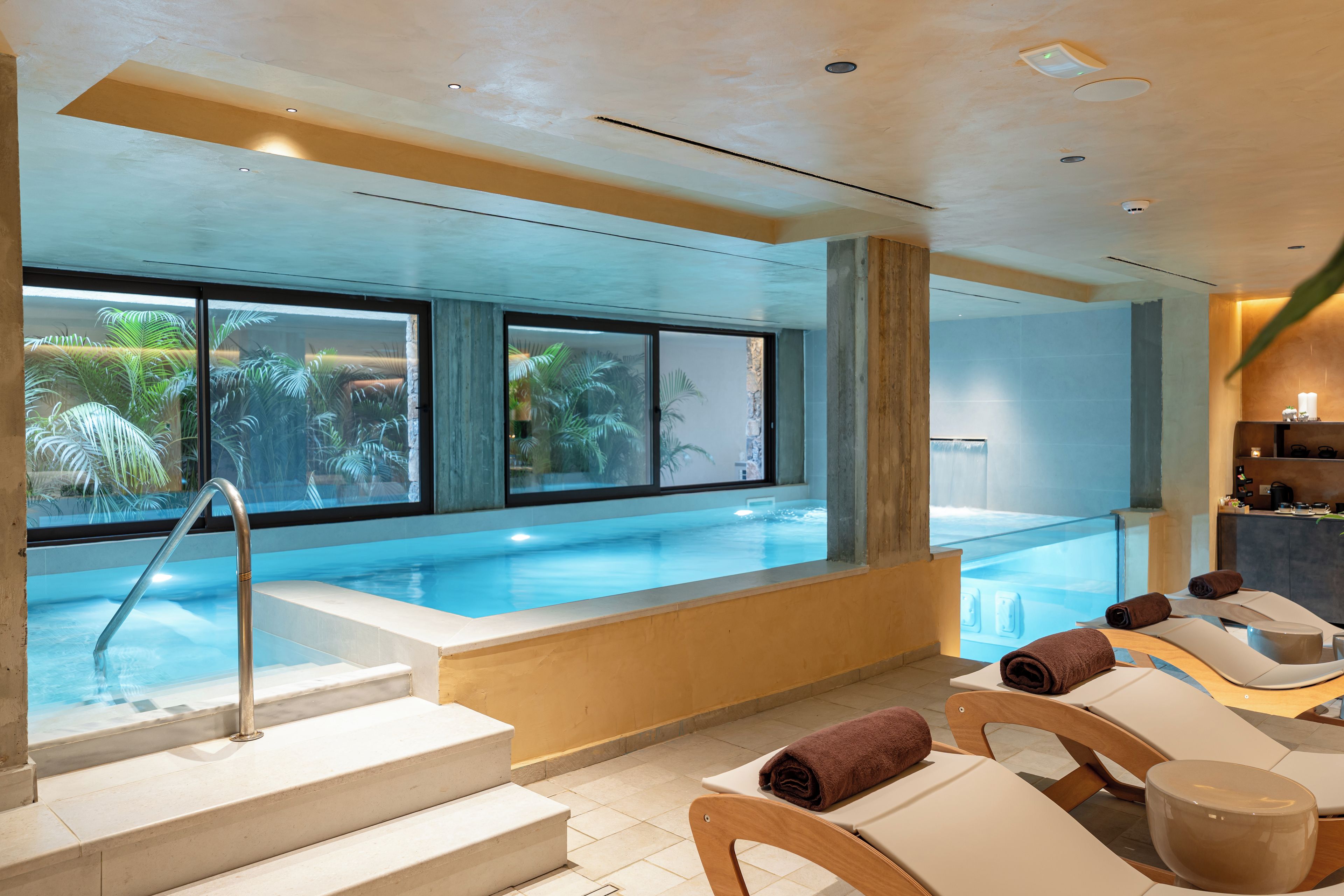 A pool area with steps leading to the water, lounge chairs, and a view of palm trees through large windows.