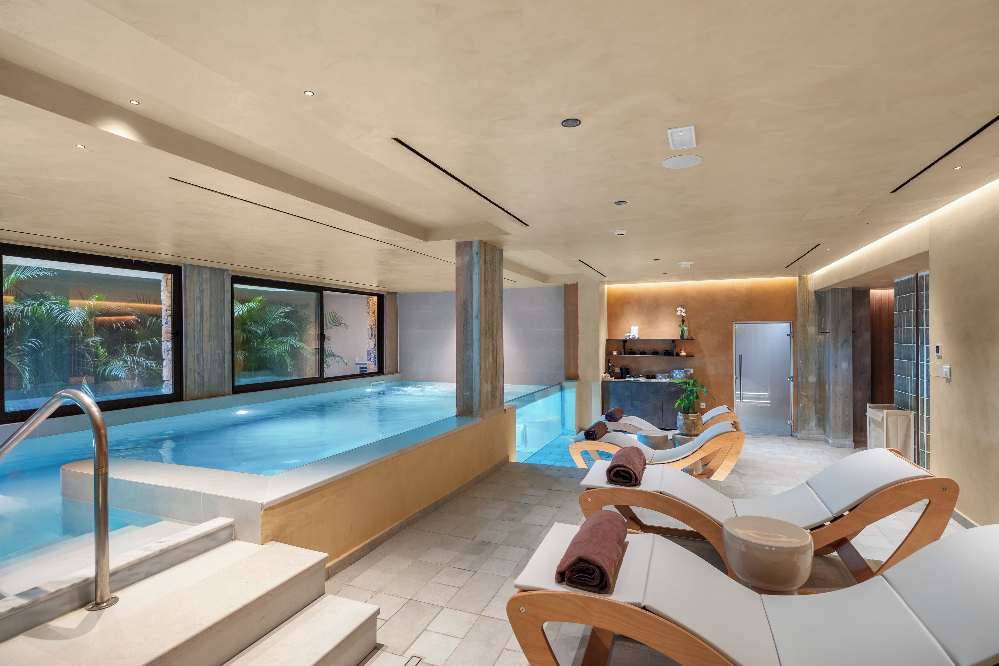 Un área de piscina interior moderna con paredes de vidrio transparente, un espacio de descanso con sillones y una cocina visible al fondo.