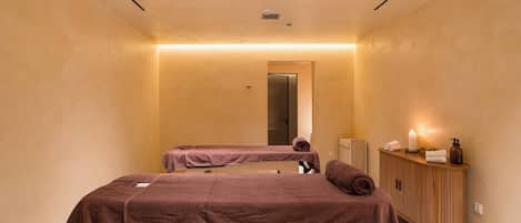 A spa room with two massage tables, a wooden cabinet with toiletries, and a mirror.