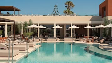 A pool area with lounge chairs and umbrellas, surrounded by a building with large windows and a palm tree in the background.