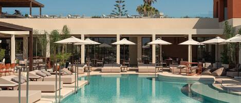 A pool area with lounge chairs and umbrellas, surrounded by a building with large windows and a palm tree in the background.