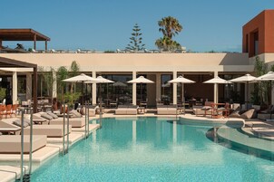 Un área de piscina con sillas de descanso y sombrillas, rodeada por un edificio con grandes ventanales y una palmera al fondo.