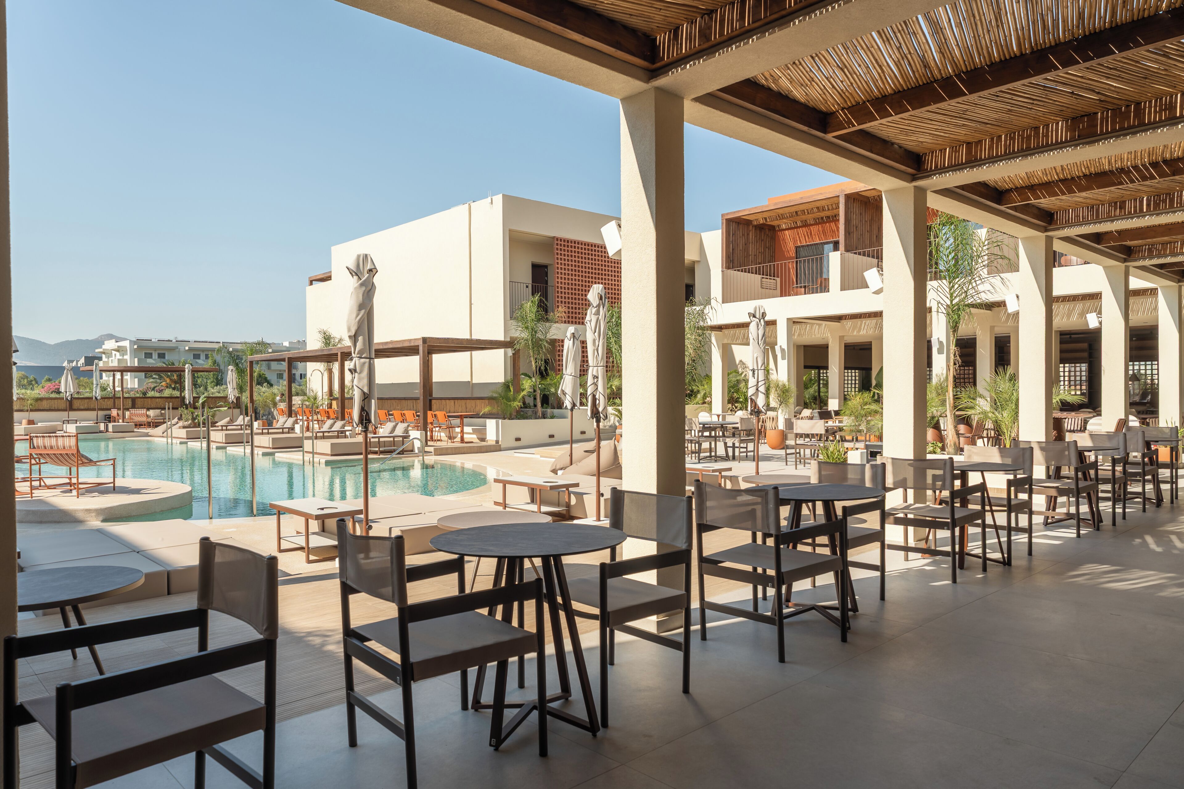 A poolside area with lounge chairs and tables, a clear sky, and buildings in the background.
