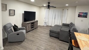 A modern living room with a gray sectional sofa, a gray recliner, a wooden dining table with chairs, a flat-screen TV, and a ceiling fan. - New! Aurora Viewing, 3 mins from airport, Prime Location (Fairbanks)