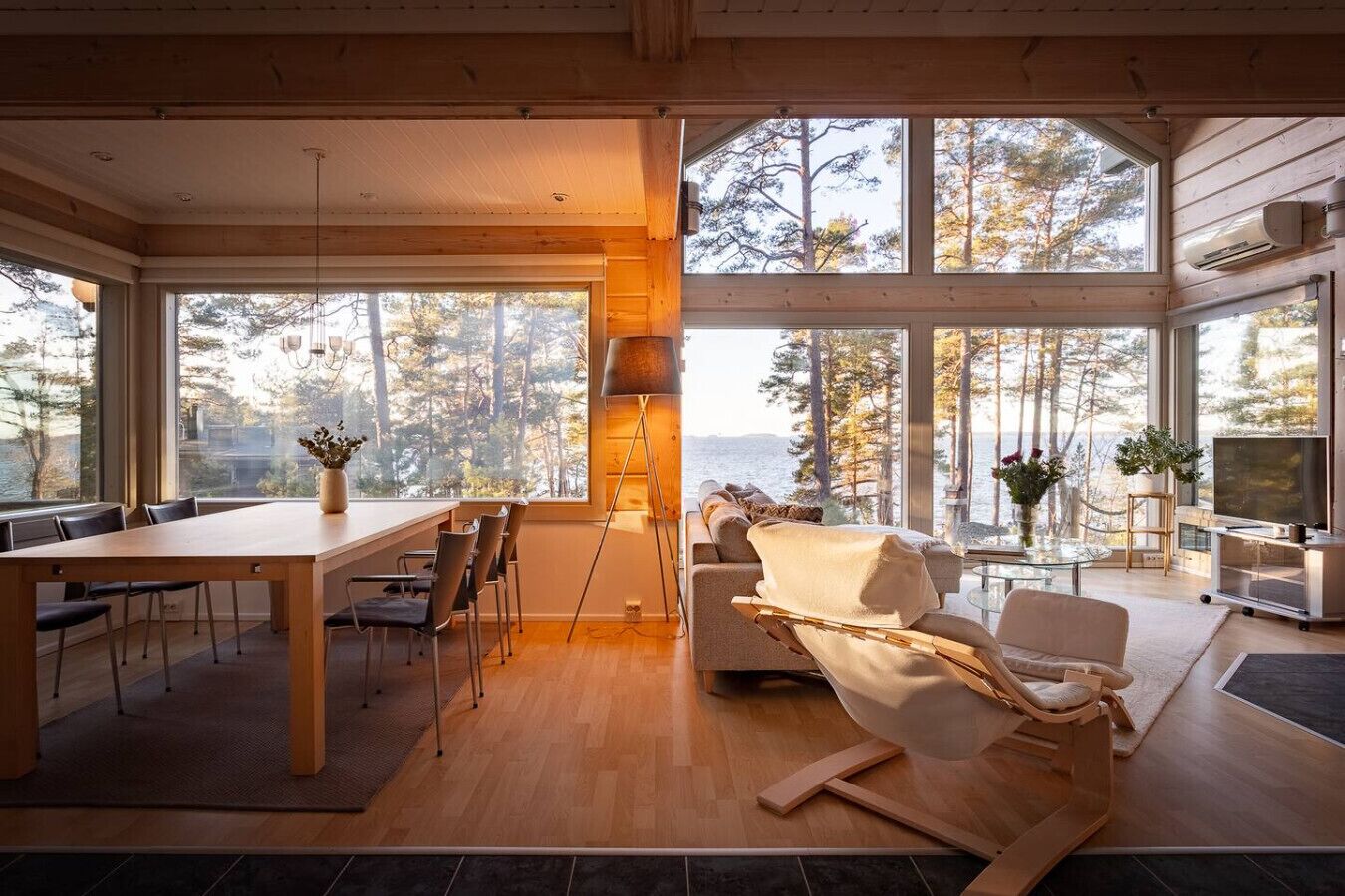 Ein großzügiger Wohnbereich mit Holzboden, einem Esstisch mit Stühlen, einer Couch und einem Fernseher. Große Fenster bieten Blick auf Bäume und ein Gewässer.