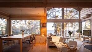 Un vaste espace de vie doté d’un plancher en bois, d’une table à manger avec des chaises, d’un canapé et d’un téléviseur. De grandes fenêtres offrent une vue sur les arbres et un plan d’eau.