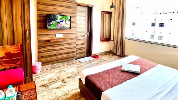 A hotel room with a bed, a desk with a chair, a television mounted on a wooden panel, and a window with a view of buildings. - Hotel Woodcrest (Zirakpur)