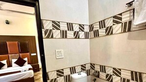 A hotel bathroom with a toilet, a sink, and a shower area. There is a bed visible through an open door in the adjacent room. - Hotel Woodcrest (Zirakpur)