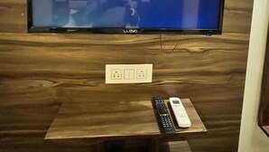 A flat-screen TV mounted on a wooden wall with a Netflix logo displayed. Below the TV, there is a small wooden table with a remote control and a wall socket. - Hotel Woodcrest (Zirakpur)