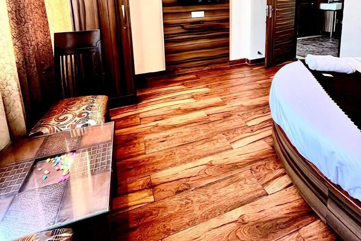Habitación de hotel con piso de madera, una cama, una mesita con mantel estampado, una silla, un televisor montado en una pared de paneles de madera y una puerta que da a un baño.