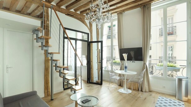 A modern living room with a staircase, a chandelier, a TV, and a balcony with a view of buildings. - Charming  Apartment Le Marais Center of Paris (Paris)