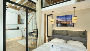 A modern bedroom with a wooden headboard, a bed with white linens, a painting of a cityscape, and a staircase leading to a kitchen area. - Charming  Apartment Le Marais Center of Paris (Paris)