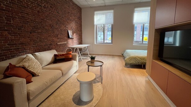 A modern living room with a brick wall, a sofa, a round coffee table, a TV, and a bed in the background.